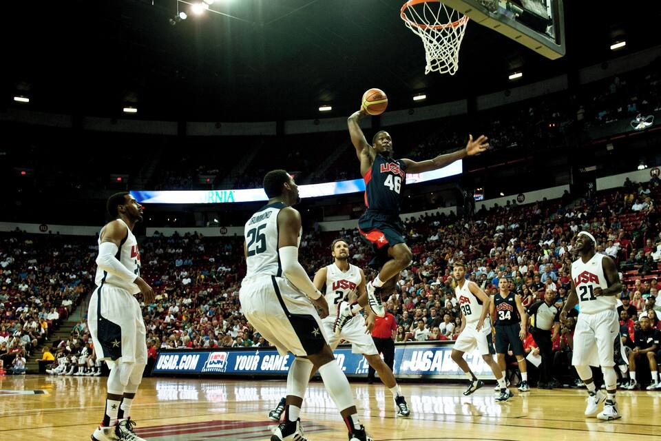 A basketball game with players in USA jerseys, a basketball hoop, and a crowd in the background.