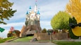 A castle with multiple towers and a flag on top, surrounded by trees and a paved area.