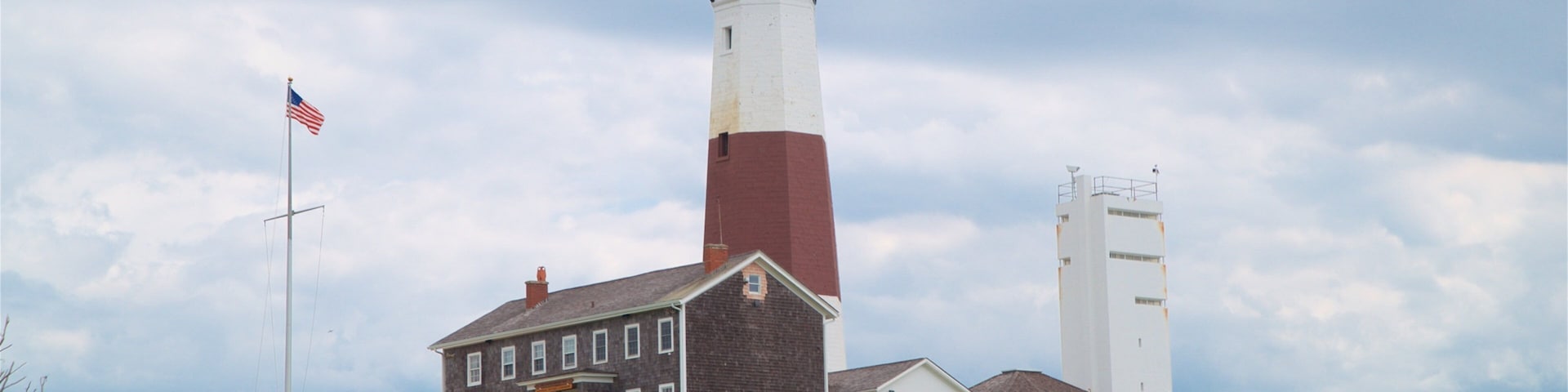 Um farol com torre vermelha e branca, um edifício branco com telhado marrom e uma bandeira americana.