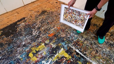 A person holding a framed painting on a floor covered with paint splatters.
