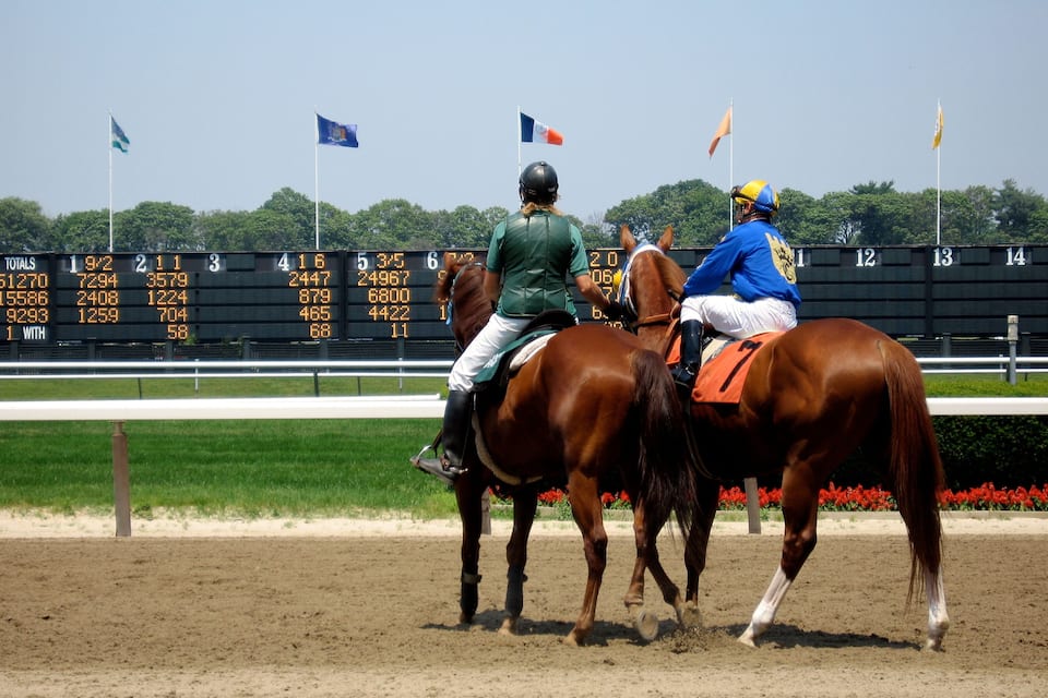 Jockeys on horses at a racetrack with a scoreboard in the background.