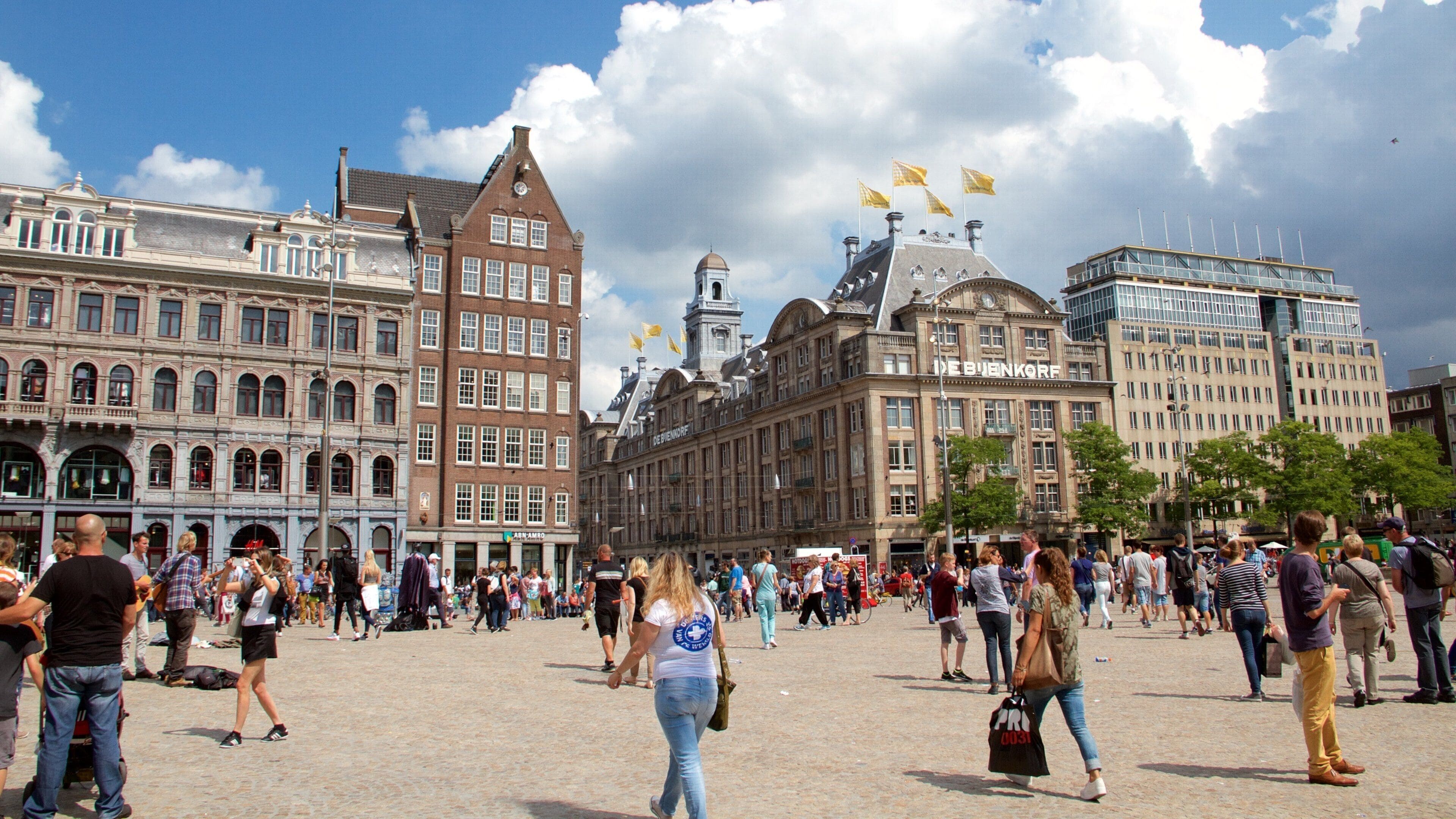 Uma praça movimentada com edifícios históricos, incluindo uma estrutura imponente de tijolos vermelhos e um edifício moderno de vidro.
