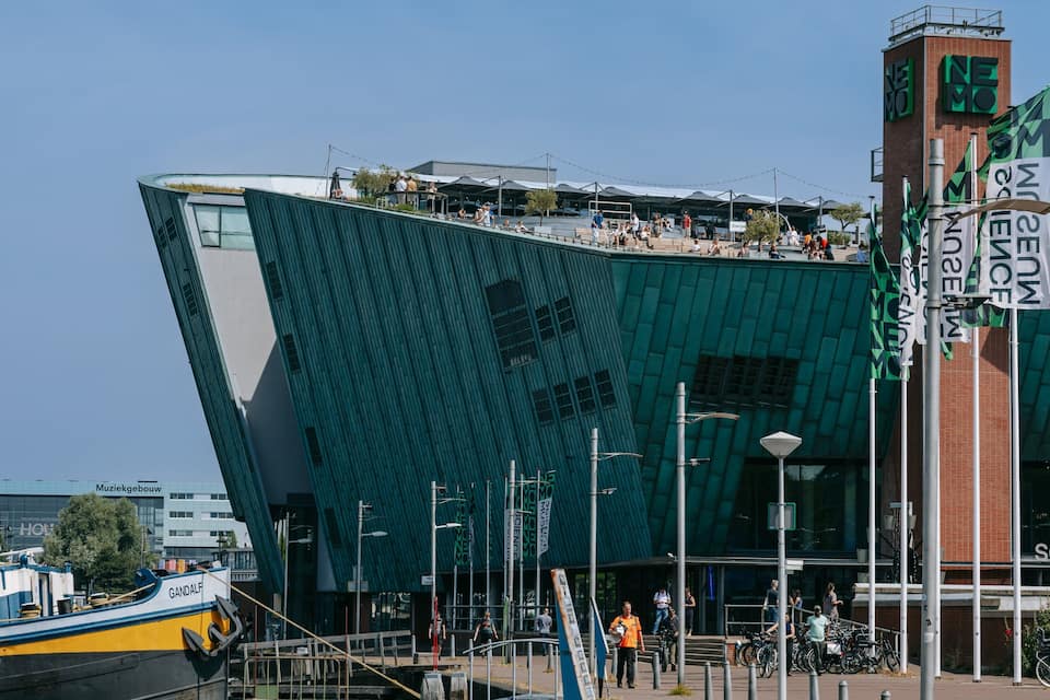 A modern building with a unique angular design, a dock with a boat, and people walking around.