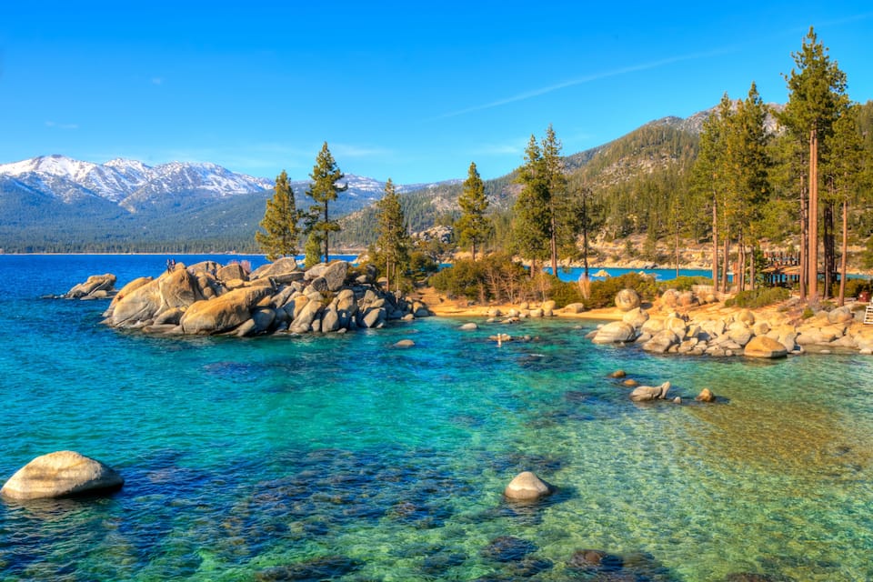 A clear lake surrounded by rocks and trees, with mountains in the background.