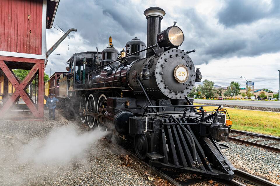 A steam locomotive numbered 25 at a station with a red building in the background.