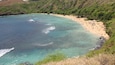 Une plage côtière aux eaux turquoise et limpides, bordée d’un rivage sablonneux et entourée de collines verdoyantes.