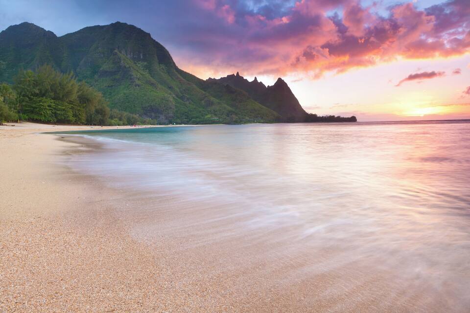 A beach with clear turquoise water, a sandy shore, and a mountain in the background.