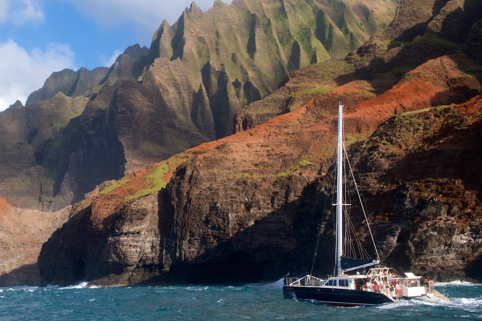 A sailboat navigating near a rocky coastline with steep cliffs and a clear sky.