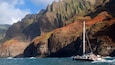 A sailboat navigating near a rocky coastline with steep cliffs and a clear sky.