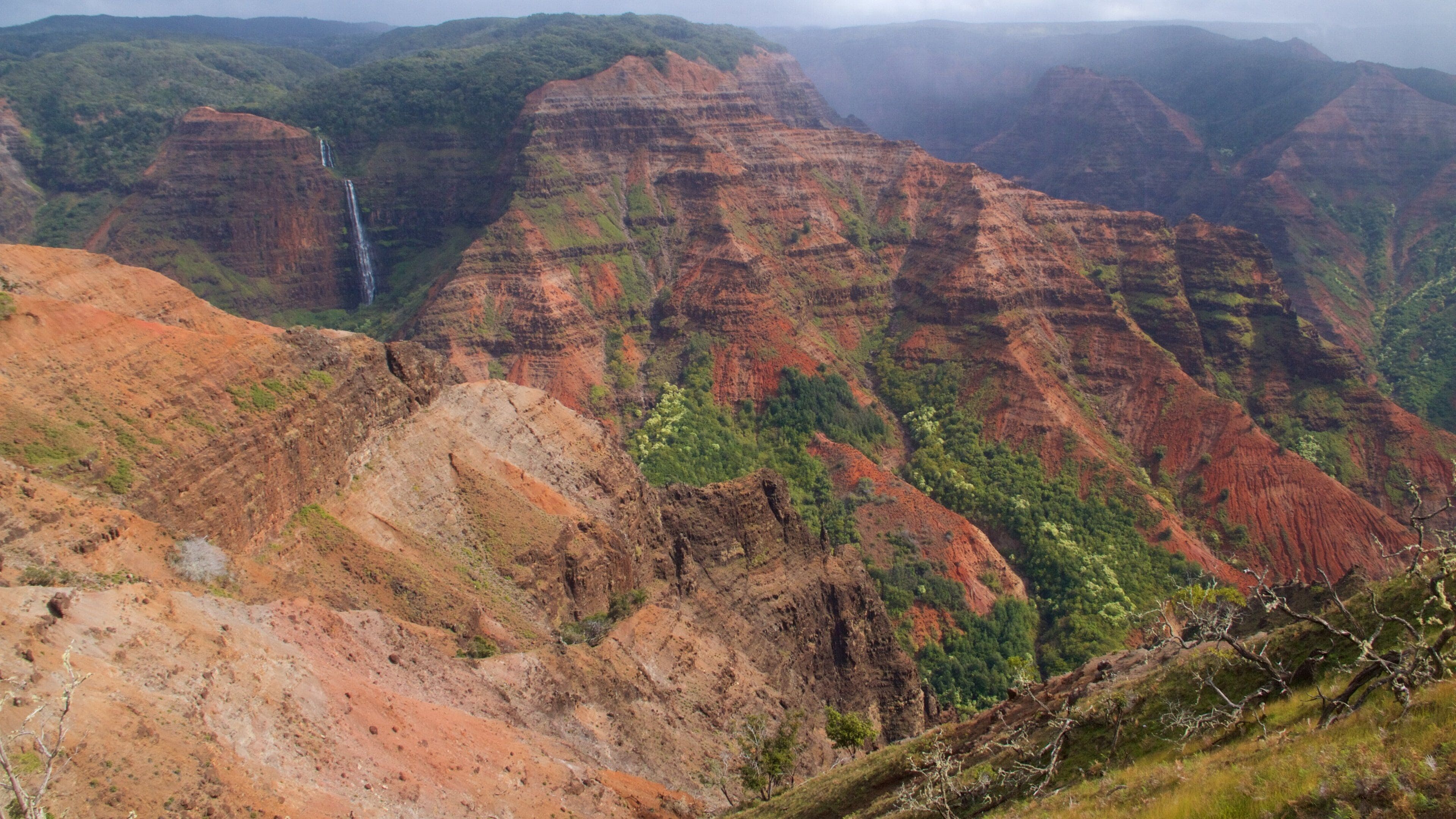 A canyon with layered red and green rock formations, a waterfall, and sparse vegetation.