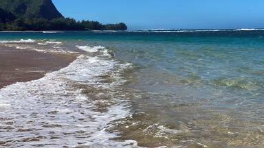 Ein Strand mit klarem Wasser, Wellen und einem Berg im Hintergrund.