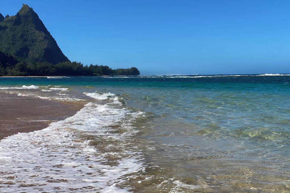 A beach with clear water, waves, and a mountain in the background.