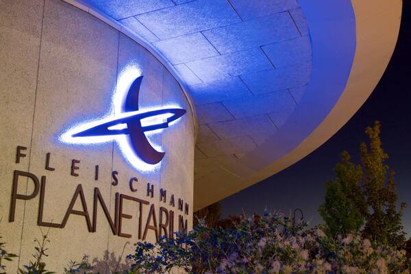 A building with a large illuminated 'x' logo and the words 'FLEISCHMANN PLANETARIUM' on its facade.