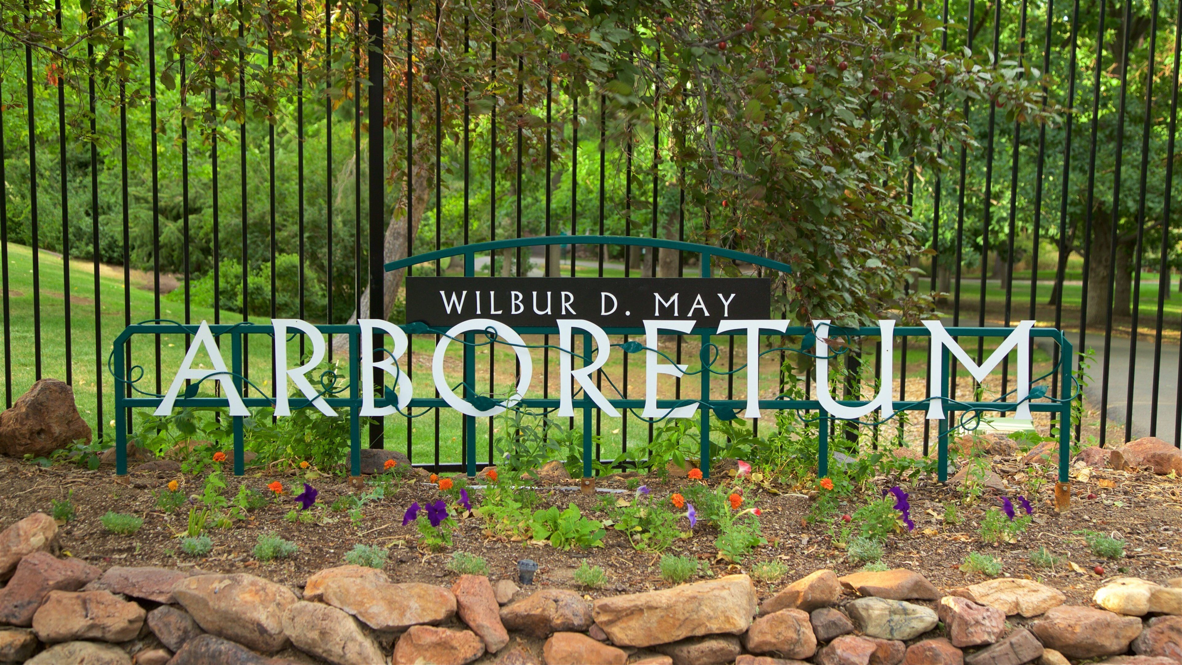 Een bord met de naam 'Wilbur D. May Arboretum', omringd door een metalen hek en bloeiende planten.