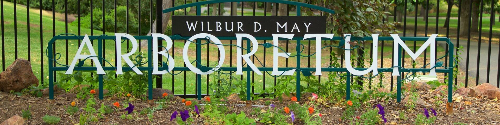Un letrero con el nombre 'Wilbur D. May Arboretum' rodeado por una cerca metálica y plantas florecientes.