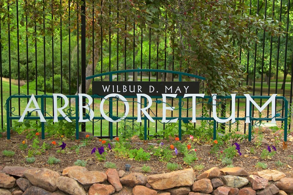 Ein Schild mit dem Namen 'Wilbur D. May Arboretum', umgeben von einem Metallzaun und blühenden Pflanzen.