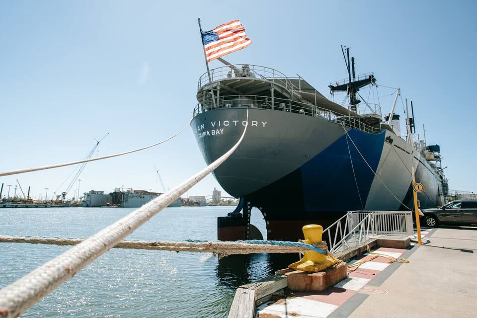 Een groot schip genaamd 'San Victory' ligt aangemeerd in een jachthaven, met de Amerikaanse vlag wapperend.