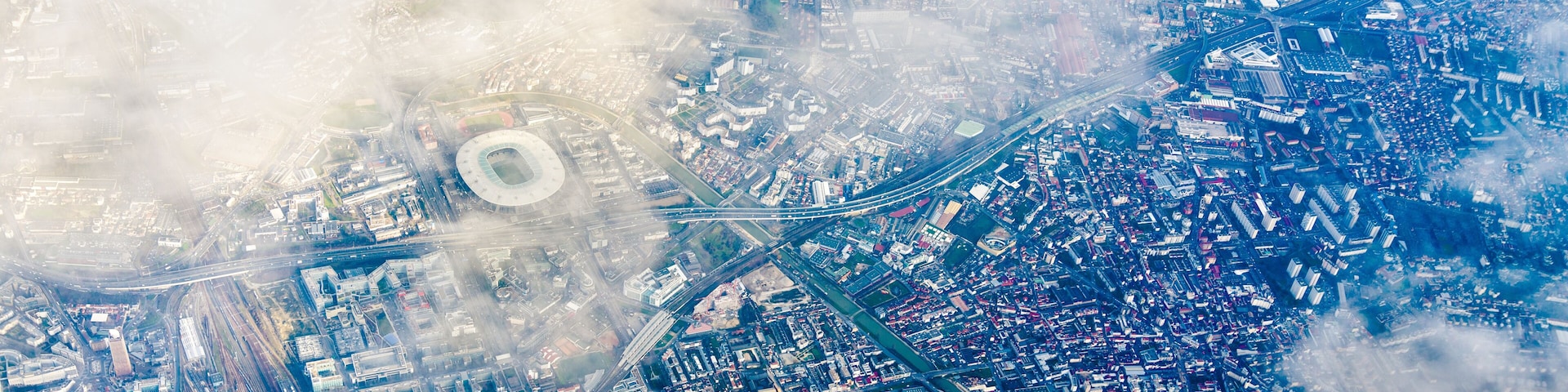 Vista aérea de uma cidade com um estádio grande, áreas urbanas densas e uma rodovia importante.