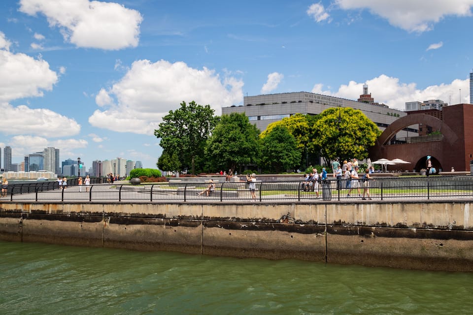 Een promenade aan het water met een modern gebouw, bomen en een rivier.