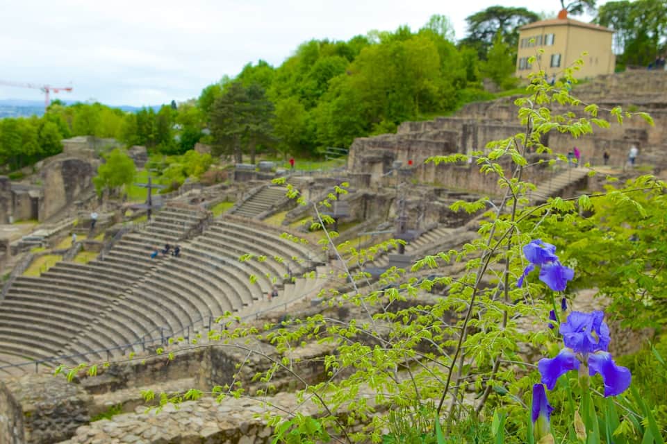 An ancient amphitheater surrounded by lush greenery and a building in the background.