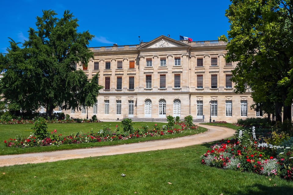 A large classical building with a symmetrical facade, surrounded by well-maintained gardens and a clear blue sky.