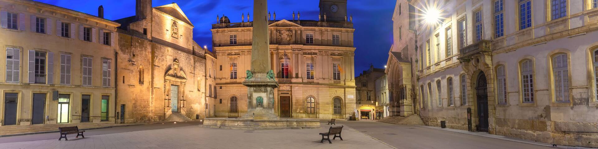 A historic square with a prominent obelisk, classical buildings, and benches.
