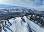 Pendiente nevada de una montaña con telesillas, pinos y cumbres nevadas a lo lejos.