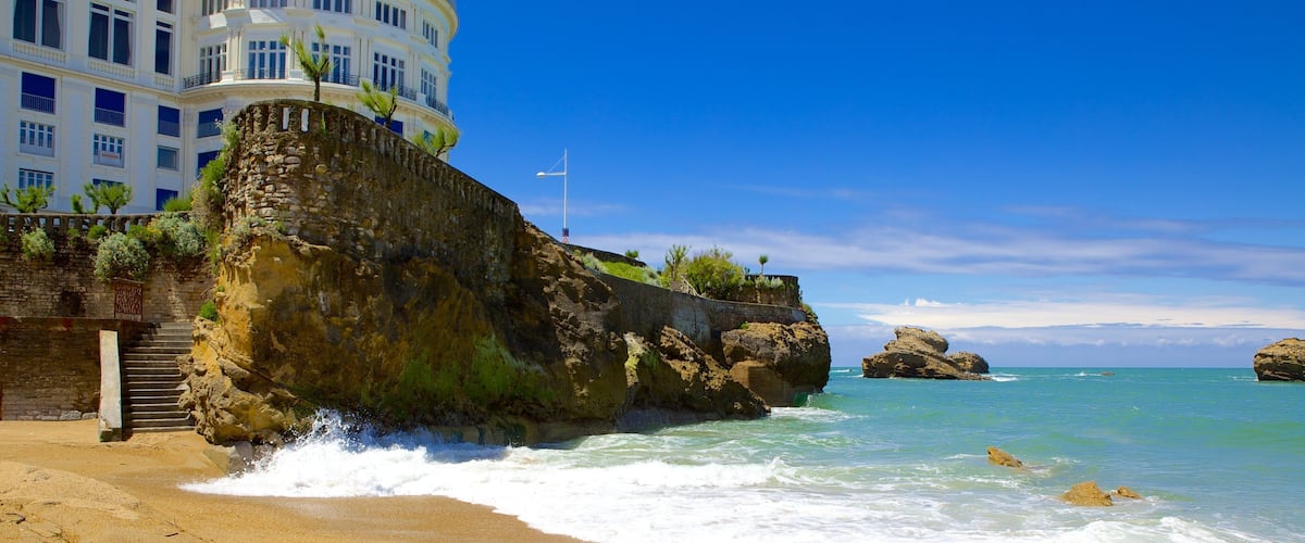 A beach with waves crashing onto the shore, a rocky cliff, and a building in the background.