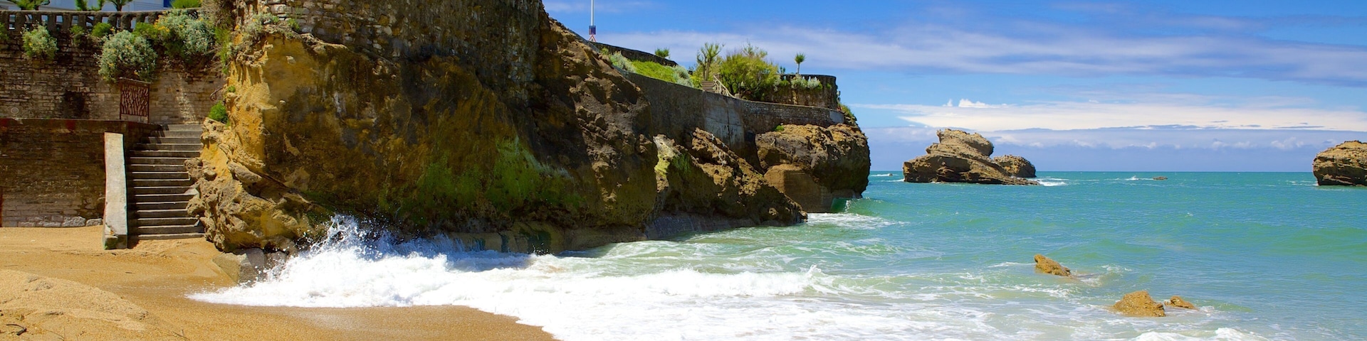 A beach with waves crashing onto the shore, a rocky cliff, and a building in the background.