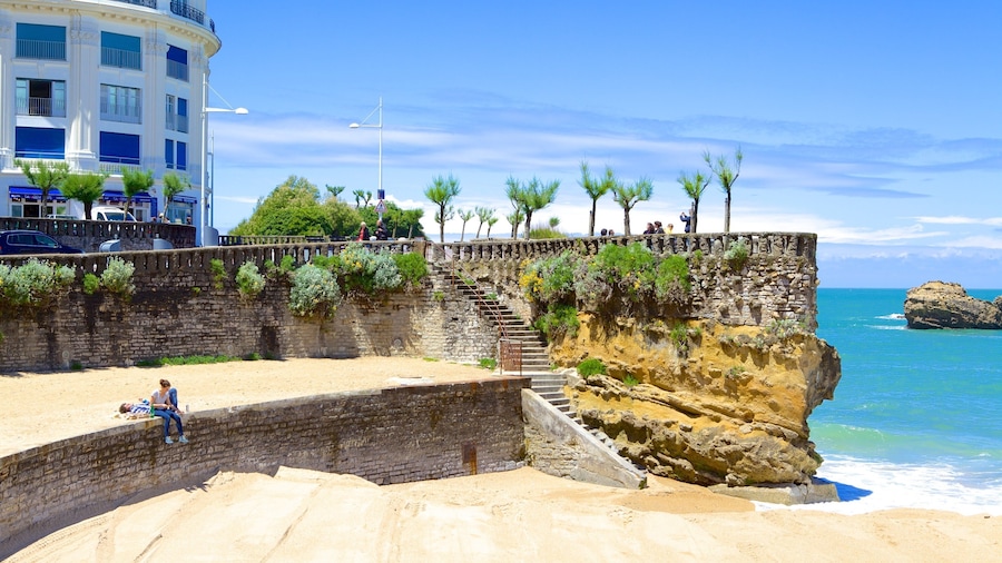 Una passeggiata costiera con un muro in pietra, scale che scendono verso la spiaggia e un edificio con balconi affacciati sul mare.