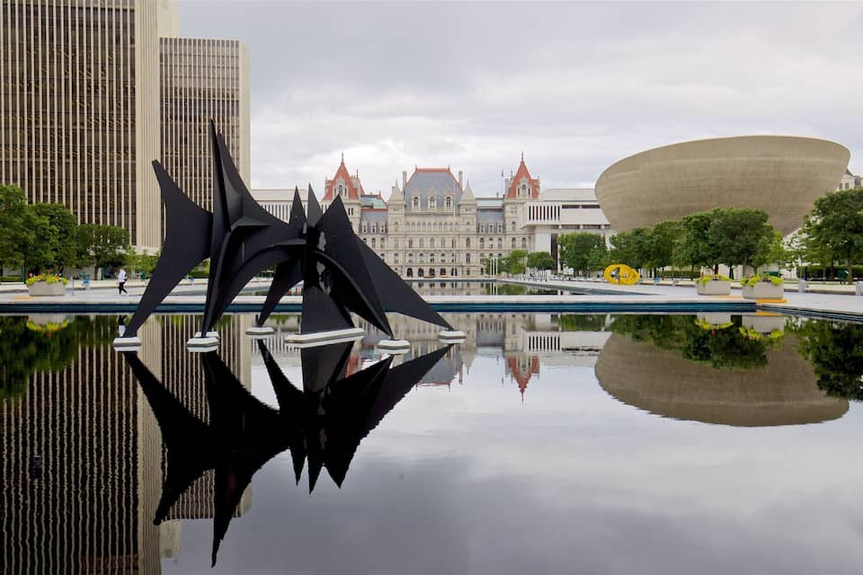 A modern sculpture in a reflective pool with historic buildings in the background.
