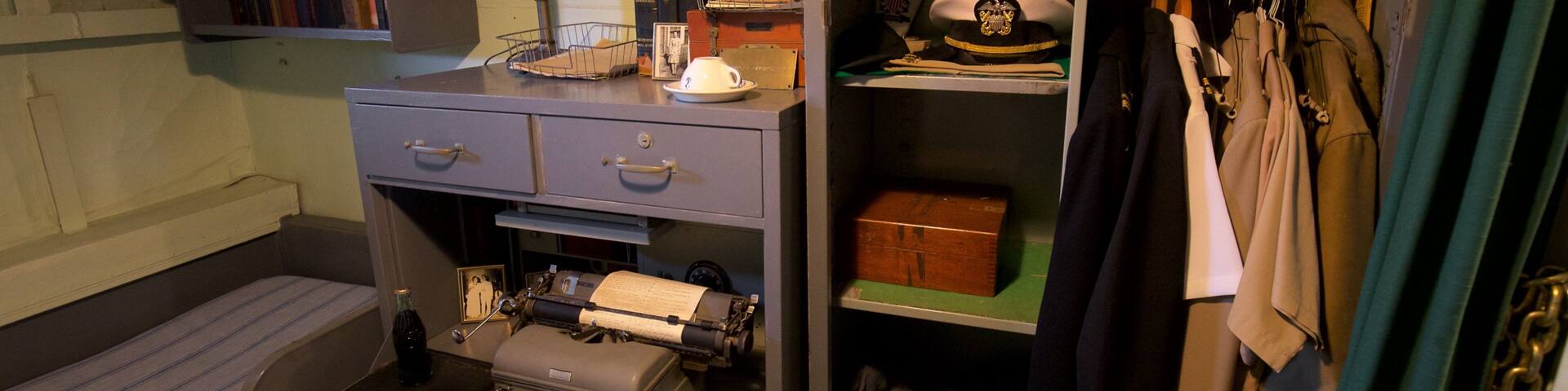 A vintage office with a typewriter, bookshelves, and a coat rack with hats.