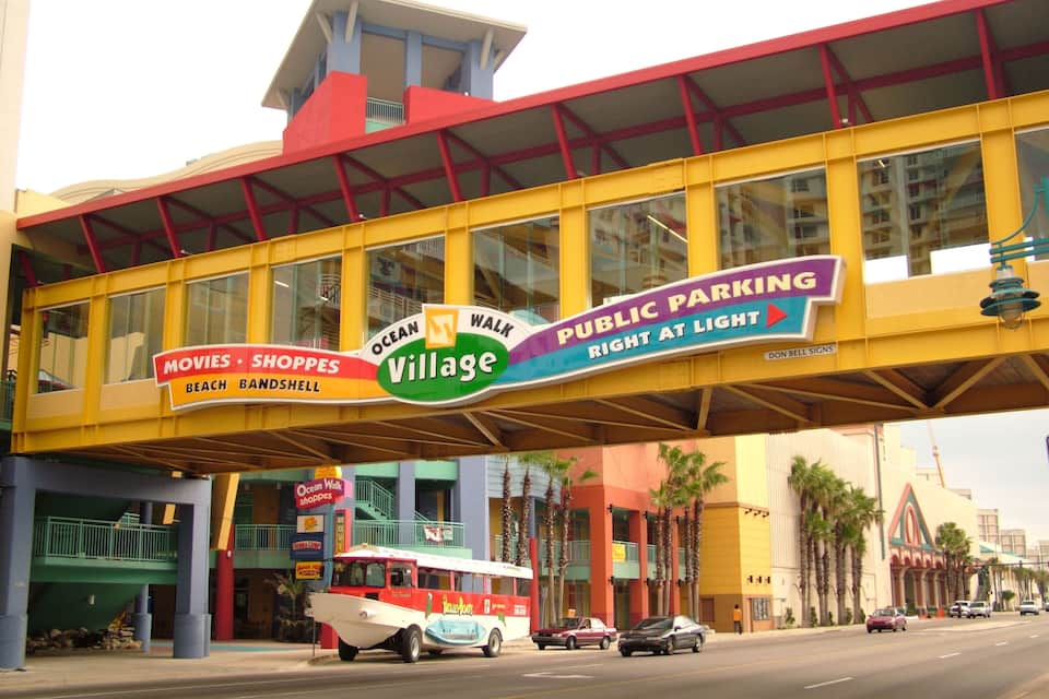 A colorful pedestrian bridge with signs for Ocean Walk Village, movies, shops, and public parking.