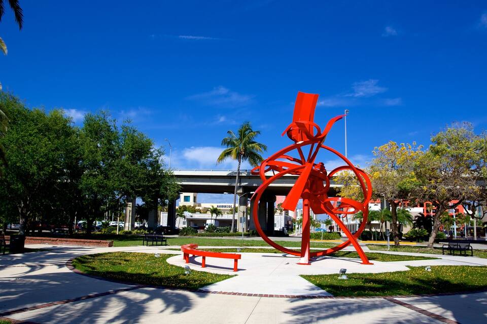 A red sculpture in a park with palm trees and a building in the background.