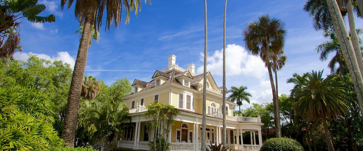 A large, yellow colonial-style house surrounded by lush greenery and tall palm trees.
