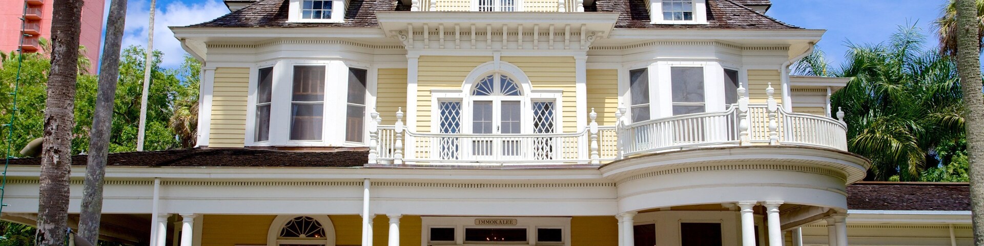 Uma grande casa de estilo vitoriano, amarela, com varanda em volta, várias janelas e um balcão.