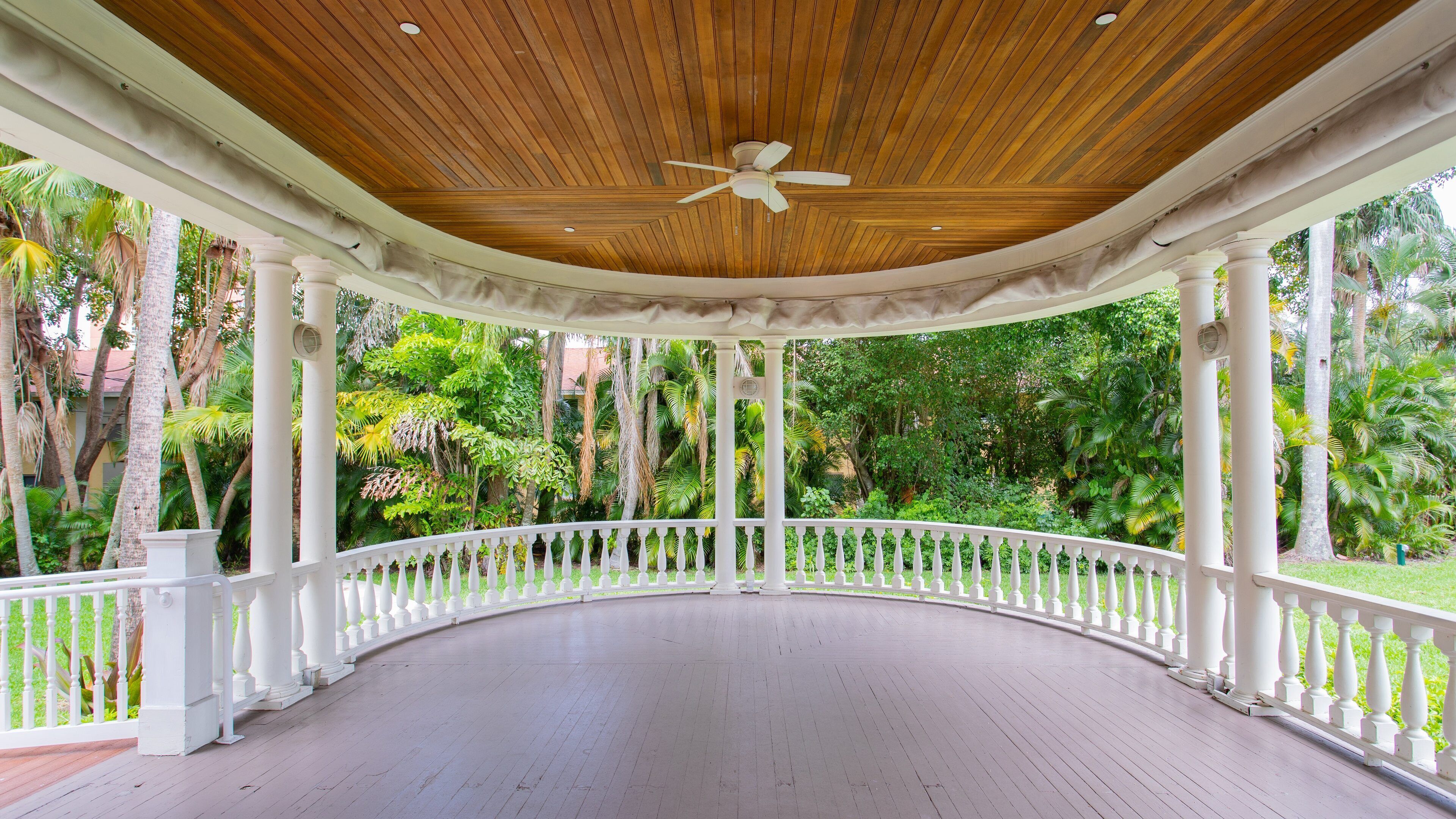 A covered wooden deck with a white balustrade, surrounded by lush tropical vegetation.