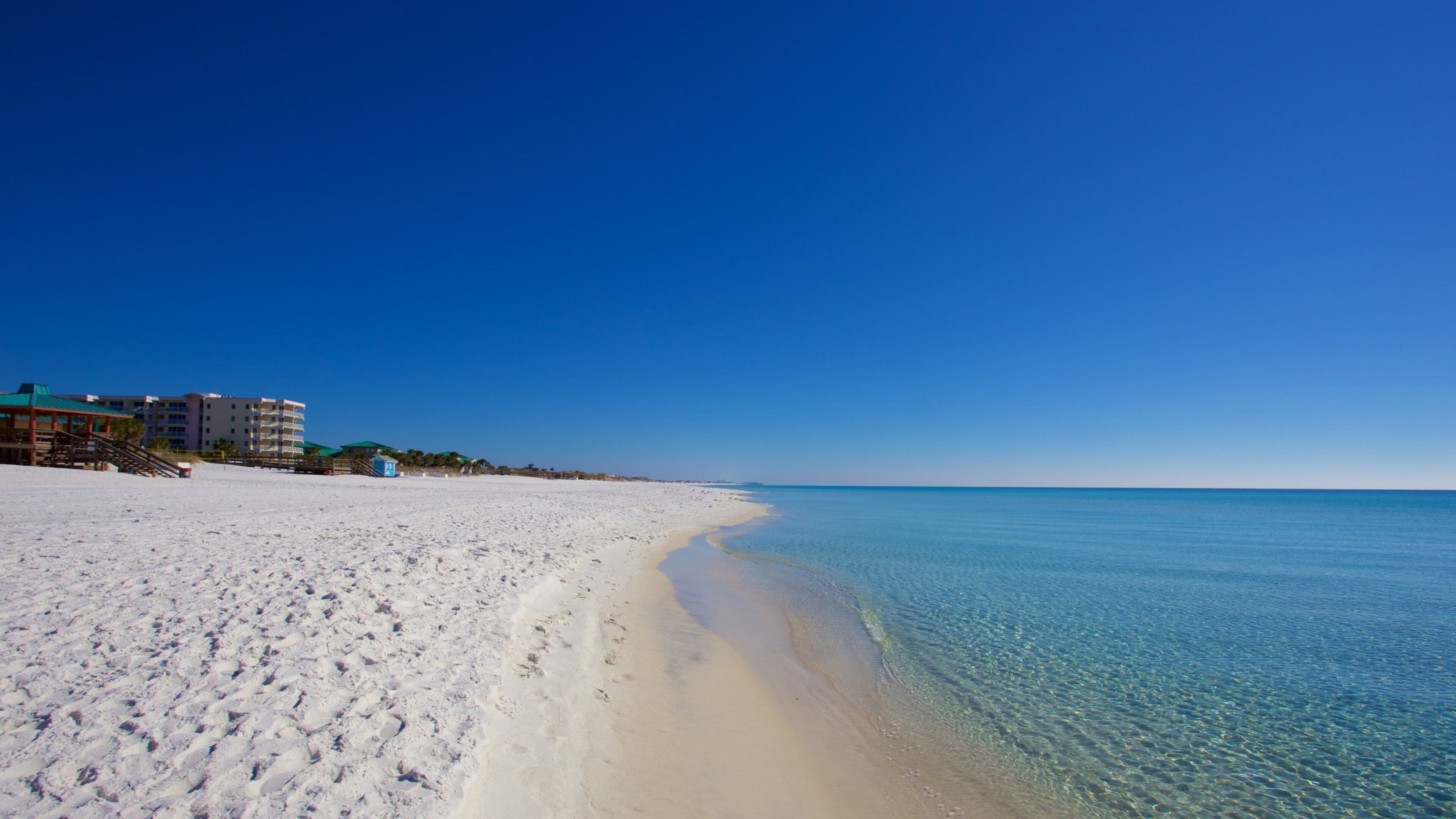 A sandy beach with clear blue water and a few buildings in the background.