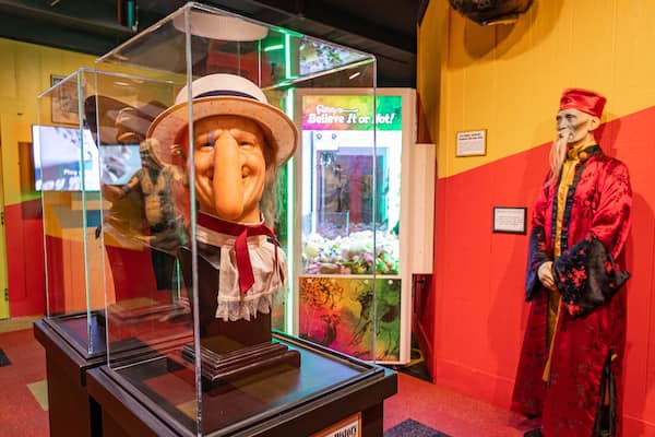 A museum exhibit with a glass display case containing a mannequin dressed in a hat and a red dress. In the background, there is a red wall with a neon sign and a statue of a man in a red coat.