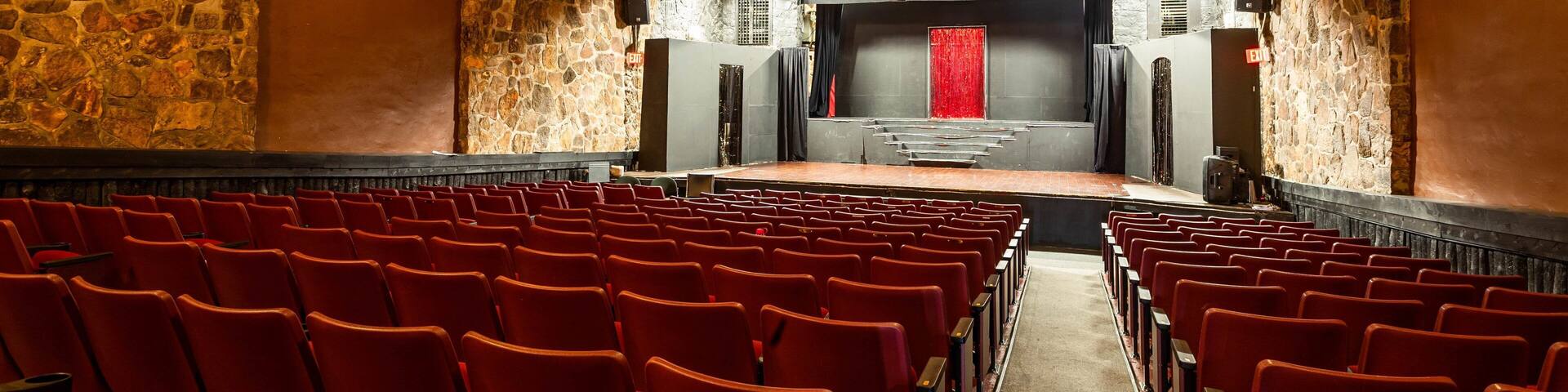 An indoor theater with red seats, a stage with a red curtain, and stone walls.