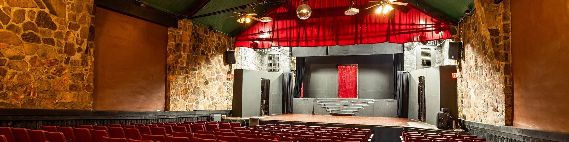Un teatro interior con asientos rojos, un escenario con cortina roja y paredes de piedra.