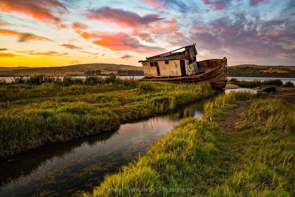 A rusted boat on a riverbank with a sunset in the background.