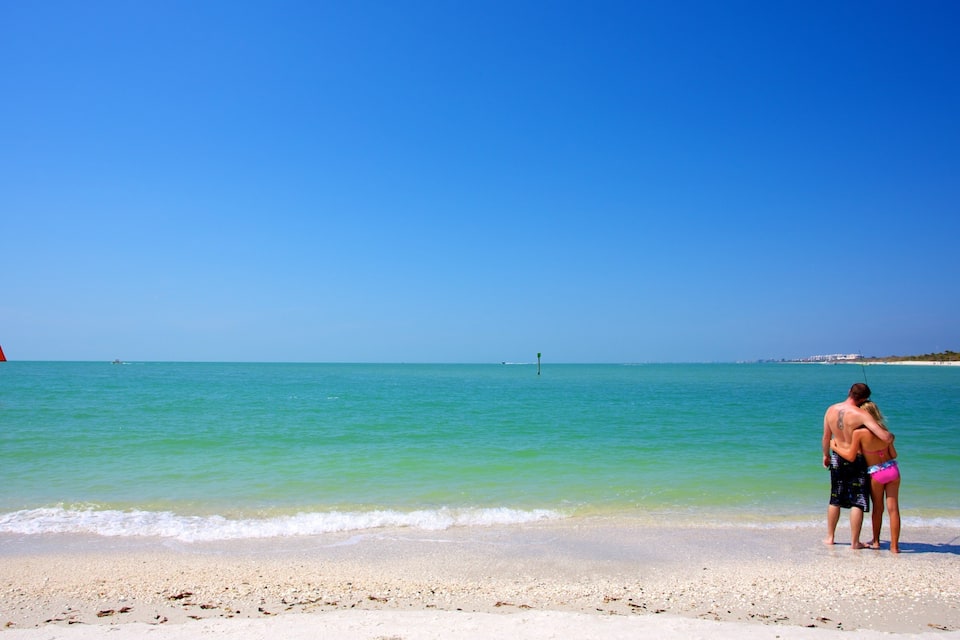 Une plage avec une eau turquoise et limpide, un rivage sablonneux, et une personne debout dans l’eau.