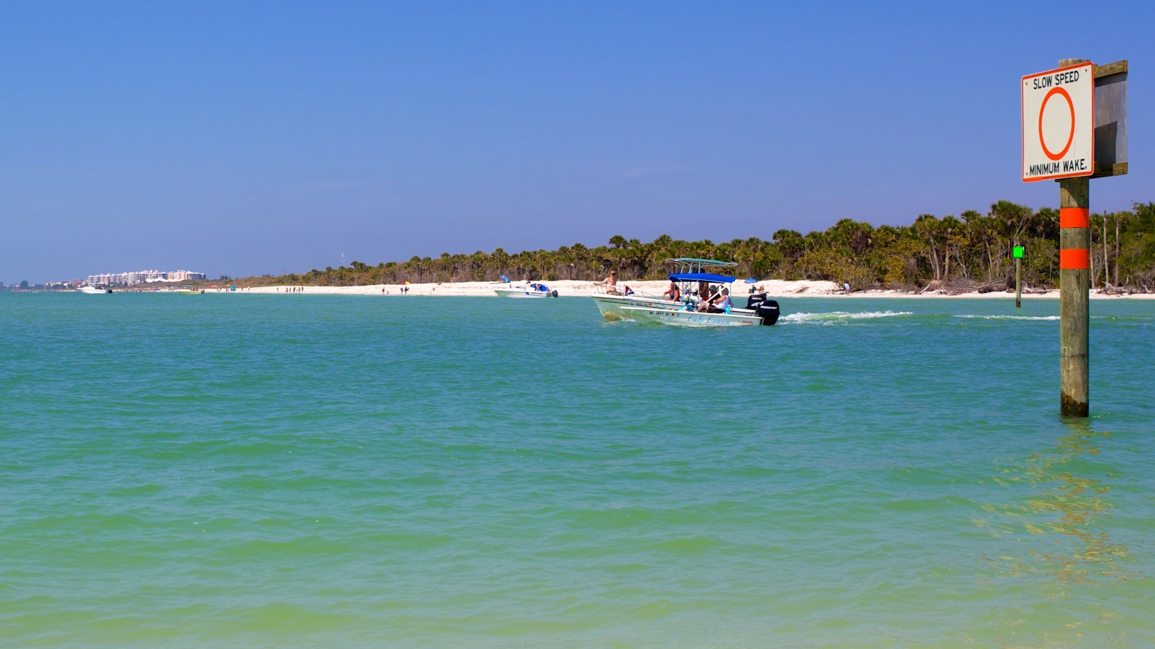 A beach with clear turquoise water, a slow speed sign, and a boat in the distance.