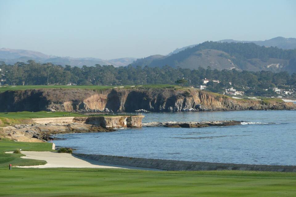 Um campo de golfe costeiro com falésias rochosas, colinas verdes e casas ao fundo.