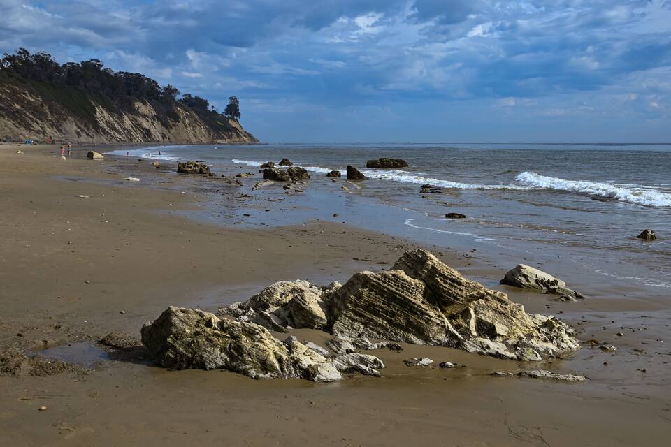 Una spiaggia sabbiosa con grandi massi in primo piano e una scogliera con alberi sullo sfondo.