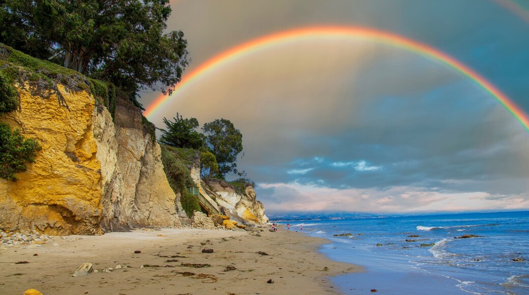 a gorgeous summer landscape at the beach with blue ocean water with waves rolling into the beach, silky brown sand and rocky cliffs covered in lush green trees and plants and blue sky and clouds