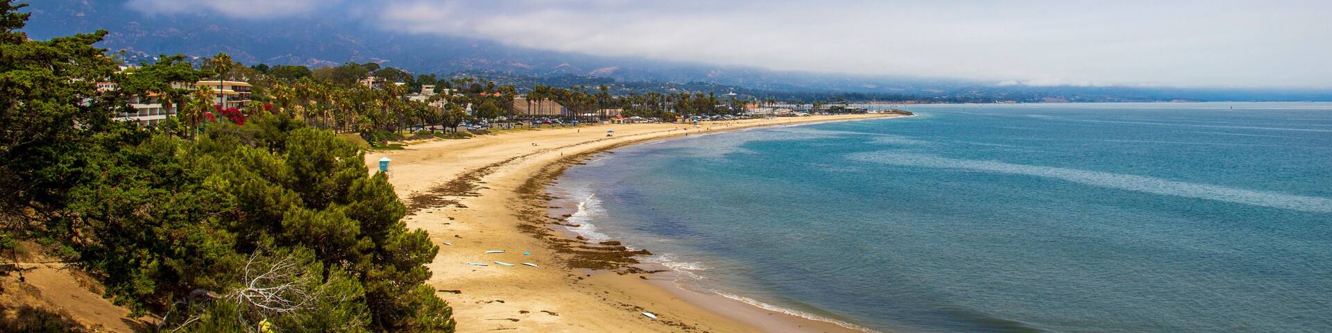 a gorgeous summer landscape at the beach with vast blue ocean water and silky brown sand surrounded by lush green trees, plants and homes with blue sky and clouds at Leadbetter Beach in Santa Barbara