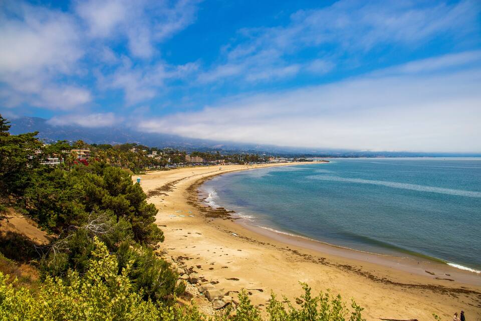 A coastal landscape with a sandy beach, dense greenery, and a clear blue sky.