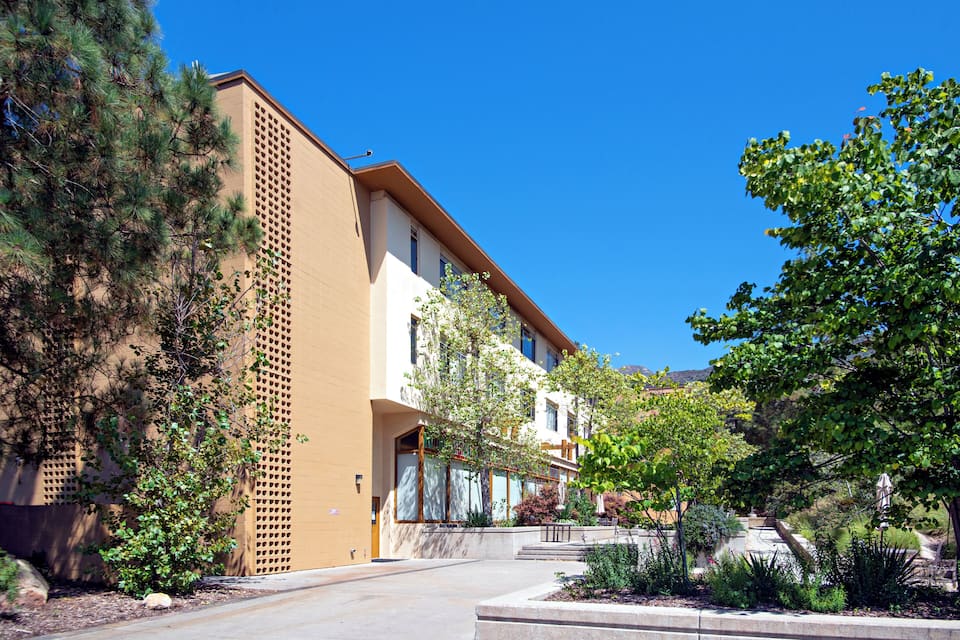 A modern building with a beige facade, surrounded by trees and a clear blue sky.